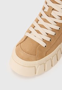 Sneaker in suede tan con lacci crema, suola chunky elevata e cuciture decorative. Presenta occhielli per i lacci e un design a punta rotonda.