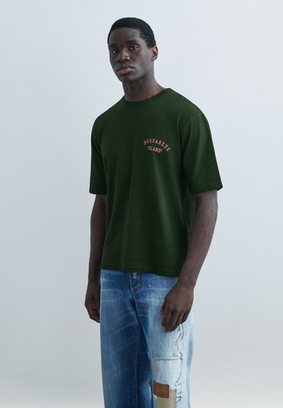 Man står i en mörkgrön Dsquared2-t-shirt och ljusblå slitna jeans med lapptäckesdetaljer på en ljusgrå bakgrund.