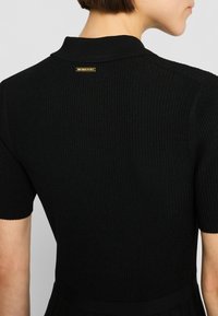 Svart ribbad stickad klänning med kort krage, korta ärmar och en figursydd överdel som övergår i en utsvängd kjol. Gulddetalj på baksidan.