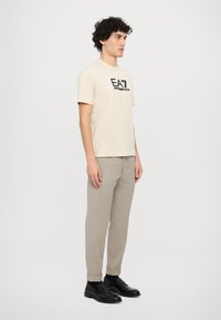 Bežové tričko s krátkým rukávem a černým logem "EA7 Emporio Armani", spárované se světle šedými chinos kalhotami a černými mokasínami.