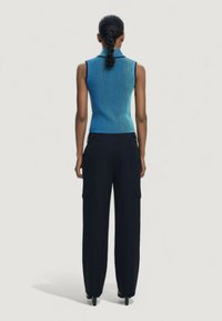 Vrouw met een gevlochten staart, gekleed in een mouwloze blauwe geribbelde top en zwarte wijde pantalon, staand met de rug naar de camera op een effen achtergrond.