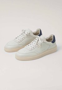 Zapatillas de cuero blancas con acentos en azul marino en el talón, puntera perforada, cordones planos y una suela de goma beige, con un diseño minimalista.