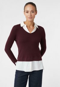 Frau mit zurückgebundenen Haaren, die einen dunkelvioletten strukturierten V-Ausschnitt-Pullover über einem weißen Hemd mit Kragen und dunklen Hosen trägt, steht mit den Händen hinter dem Rücken.