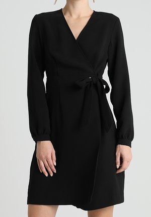 Femme portant une robe portefeuille noire à manches longues, nouée à la taille, debout devant un fond uni gris clair.