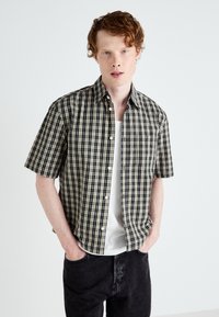 Kortærmet ternet skjorte i sort og beige med knaplukning, båret over en hvid tanktop. Bemærkelsesværdig forlommet detalje.