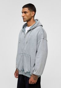 Lichtgrijze ritshoodie gemaakt van zacht materiaal, met een grote voorkantzak, een capuchon met trekkoord en geribde manchetten. Casual oversized pasvorm.