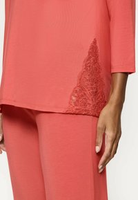 Ensemble de loungewear en corail, coupe décontractée, avec un détail en dentelle sur le côté gauche du haut. Texture douce, manches 3/4 et pantalon assorti.