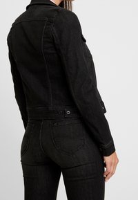Černá džínová bunda s přiléhavým střihem, klasickým límcem a knoflíkovým zapínáním. Disponuje jednou náprsní kapsou a jemnými švy jako akcenty.