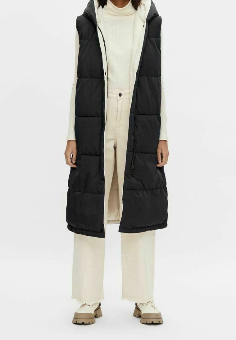 Černá prošívaná vesta s kapucí, s celým zipem, bočními kapsami a dlouhým střihem, nošená přes krémový rolák a světlé kalhoty.
