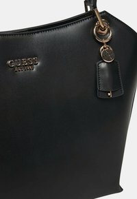 Borsa a tracolla nera in pelle con una texture liscia, caratterizzata da un logo in argento, dettagli in ottone dorato e un ciondolo removibile attaccato a un anello.