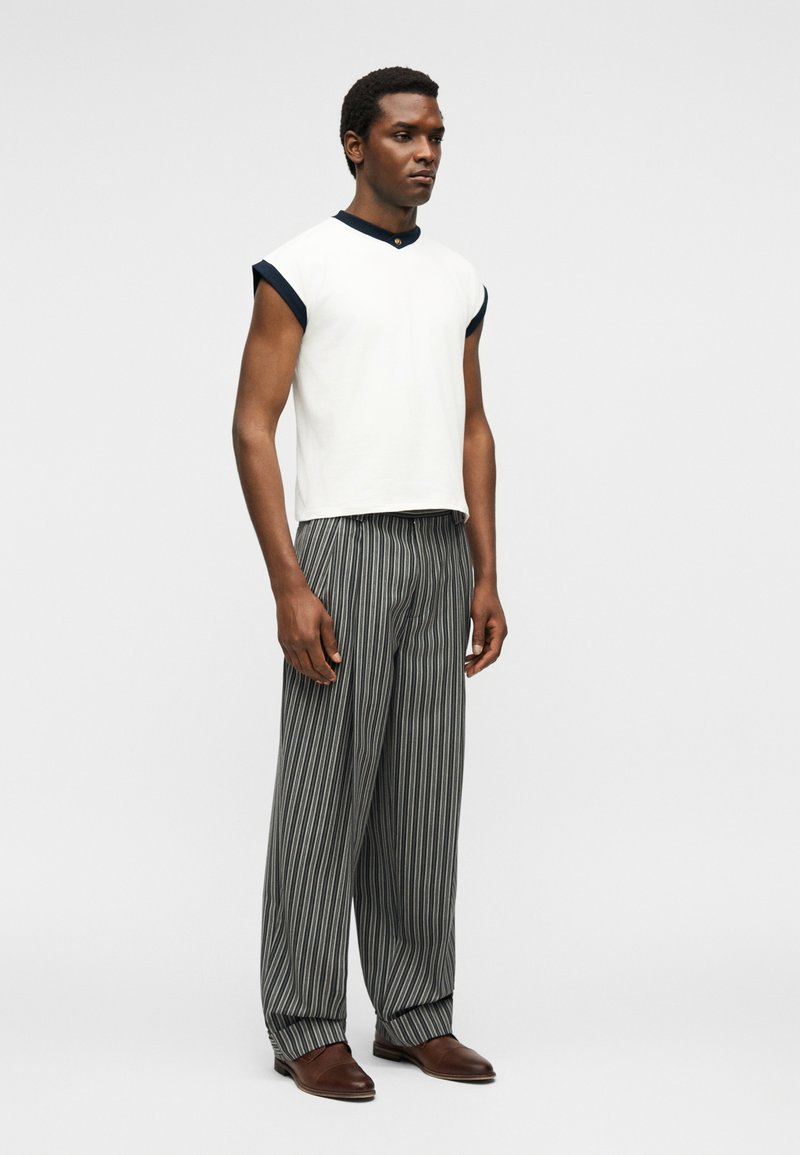 Man wearing a sleeveless white top with black trim, loose gray and black striped pants, and brown dress shoes, standing against a plain background.