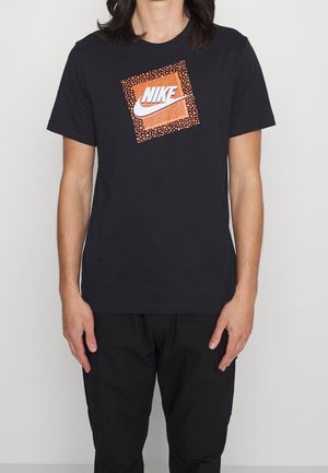 Person iført sort Nike T-shirt med orange prikket firkant og hvid Nike-logo centreret på brystet, kombineret med sorte bukser.