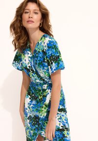 Sukienka w kwiaty z kopertowym krojem i krótkimi, falbaniastymi rękawami. Materiał zdobi niebiesko-zielony wzór akwarelowy na białym tle.