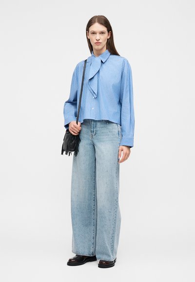 Γυναίκα που φορά μπλούζα με μπλε ρίγες και φιόγκο και τζιν παντελόνι με φαρδιά πόδια και διακοσμητικά στοιχεία, κρατώντας μια μαύρη τσάντα ώμου με φούντες, στέκεται σε λευκό φόντο.