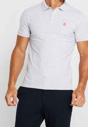 Mężczyzna w jasnoszarej koszulce polo z krótkim rękawem i małym czerwonym logo na klatce piersiowej oraz czarnych spodniach, stojący na jednolitym białym tle.