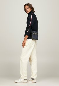 Giacca a zip navy con strisce bianche e rosse, pantaloni a gamba larga crema e sneakers crema con dettagli neri. Piccola borsa a tracolla nera.