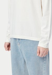 Weißes Langarmhemd mit einfachem Saum, kombiniert mit hellblauen Jeans. Der Stoff wirkt weich mit einer glatten Textur.