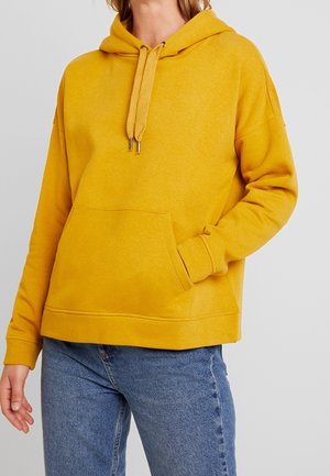 Personne portant un sweat à capuche jaune moutarde avec poche avant et cordons, associé à un jean en denim bleu, sur un fond gris uni.