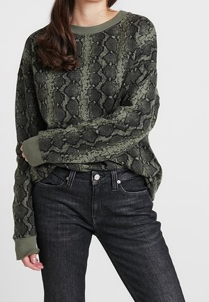 Femme portant un pull à imprimé serpent vert olive et noir avec des poignets olive et un jean noir taille haute, sur un fond blanc.