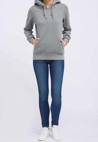 Sudadera gris con cordón, bolsillos frontales y logo. Combinada con jeans ajustados azules y zapatillas blancas. Tejido suave y corte relajado.