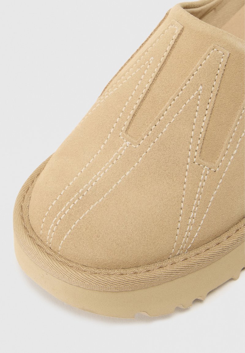 Beige suède instap schoen met opvallende stiksels, ronde neus en een gestructureerde zool met een golfpatroon.