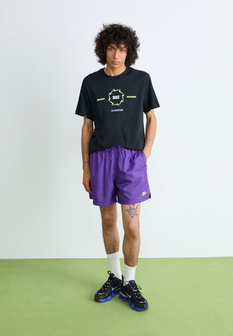 Jong persoon met krullend haar, gekleed in een zwart Nike T-shirt, paarse Nike shorts, witte sokken en zwart-blauwe sneakers, staand op een groene vloer.