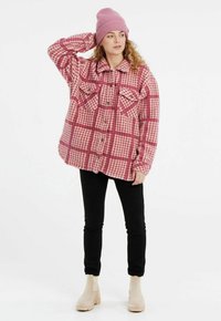 Camicia di flanella a quadri rosa con tasche frontali, chiusura con bottoni e una vestibilità rilassata, abbinata a pantaloni neri e stivali alla caviglia beige chiaro.