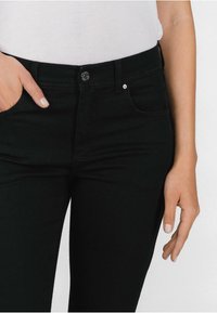 Črne kavbojke s prilegajočo se krojem, z zaponko na gumbe spredaj, dvema sprednjima žepoma in mehkim, teksturiranim materialom. Roka je vstavljena v žep.