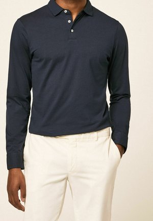Homme portant un polo à manches longues bleu marine foncé avec des boutons blancs et un pantalon beige clair, une main dans la poche, sur un fond clair.