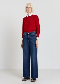 Roter gestrickter Pullover mit einem strukturierten Muster, kombiniert mit weit geschnittenen, dunkelblauen Jeans. Der Look umfasst dunkle Schuhe und ein weißes Hemd mit Kragen darunter.