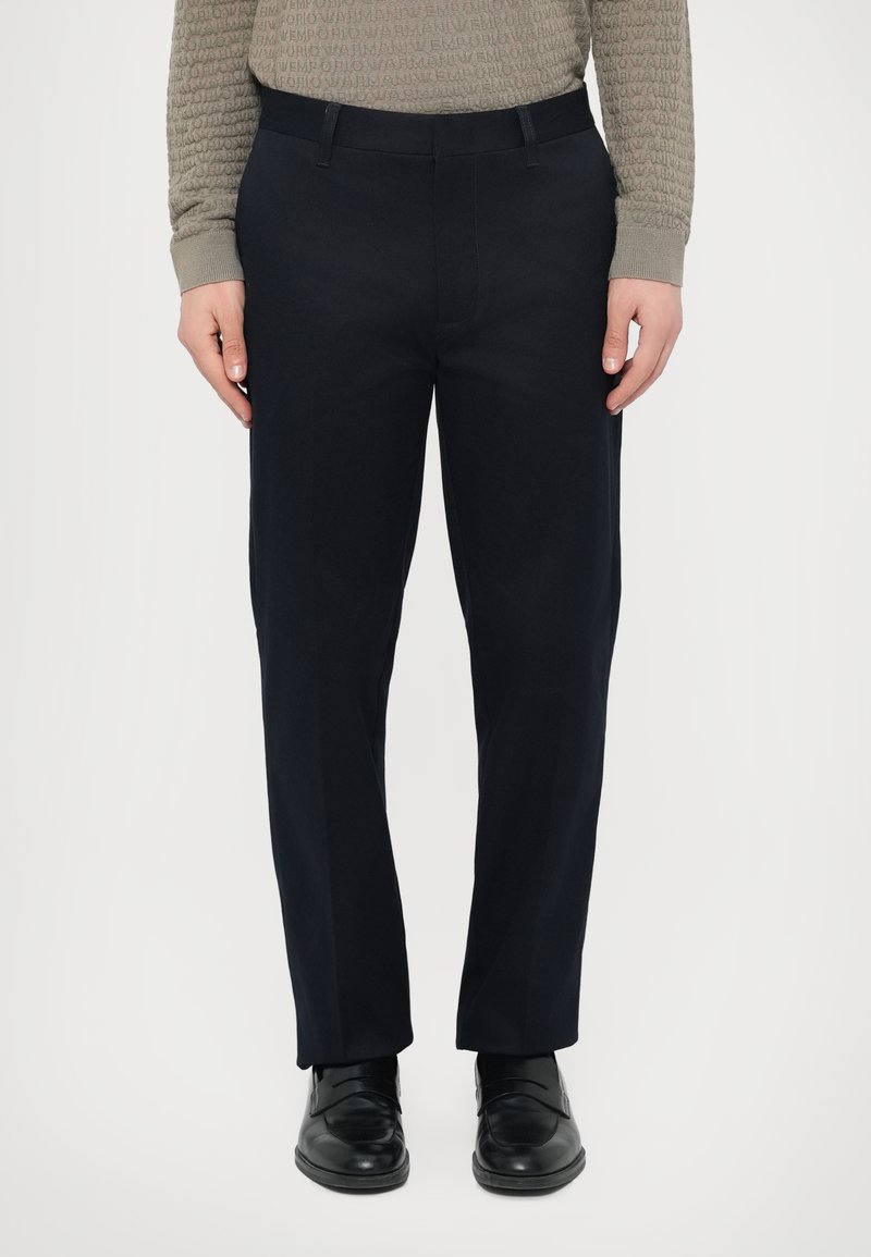 Osoba ubrana w czarne formalne spodnie, czarne skórzane buty oraz jasnobrązową, teksturowaną koszulę z długim rękawem, stojąca na białym tle.