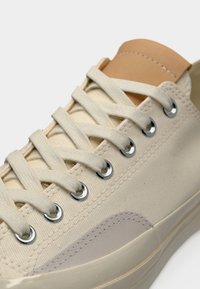 Μπεζ πάνινο αθλητικό παπούτσι με λείο δερμάτινο στοιχείο, μεταλλικά ρουλεμάν και επίπεδες κορδόνια, με στρογγυλεμένη μύτη και υφασμάτινη λεπτομέρεια στο πάνω μέρος.