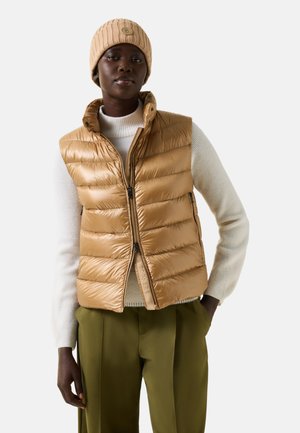 Vrouw met een beige gebreide muts en een lichtbruine puffer-vest over een witte trui met olijfgroene broek, staand met één hand in de zak.