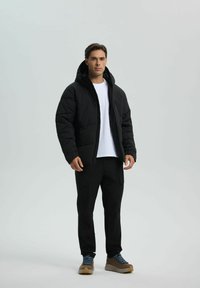 Giacca a vento nera con cappuccio e texture trapuntata, indossata sopra una t-shirt bianca. Abbinata a pantaloni cargo neri e scarpe marroni.