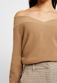 Beżowy sweter z dzianiny z odkrytymi ramionami i prążkowaną teksturą, z szerokim dekoltem. Do tego jasne spodnie w kratę.