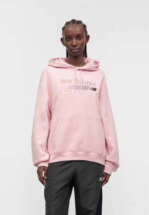 Joven con el pelo trenzado, vestida con una sudadera con capucha rosa claro de New Balance y pantalones deportivos negros, de pie frente a un fondo liso.