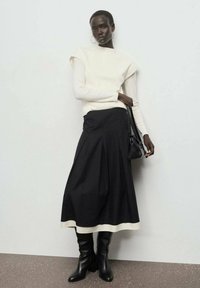 Haut crème à manches longues avec épaules structurées, associé à une jupe plissée noire et des bottes à talons noirs montantes. Sac à main noir à la main.