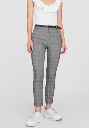 Vrouw draagt grijze geruite slim-fit broek met zwarte riem, wit mouwloos topje met ruches en witte chunky sneakers.