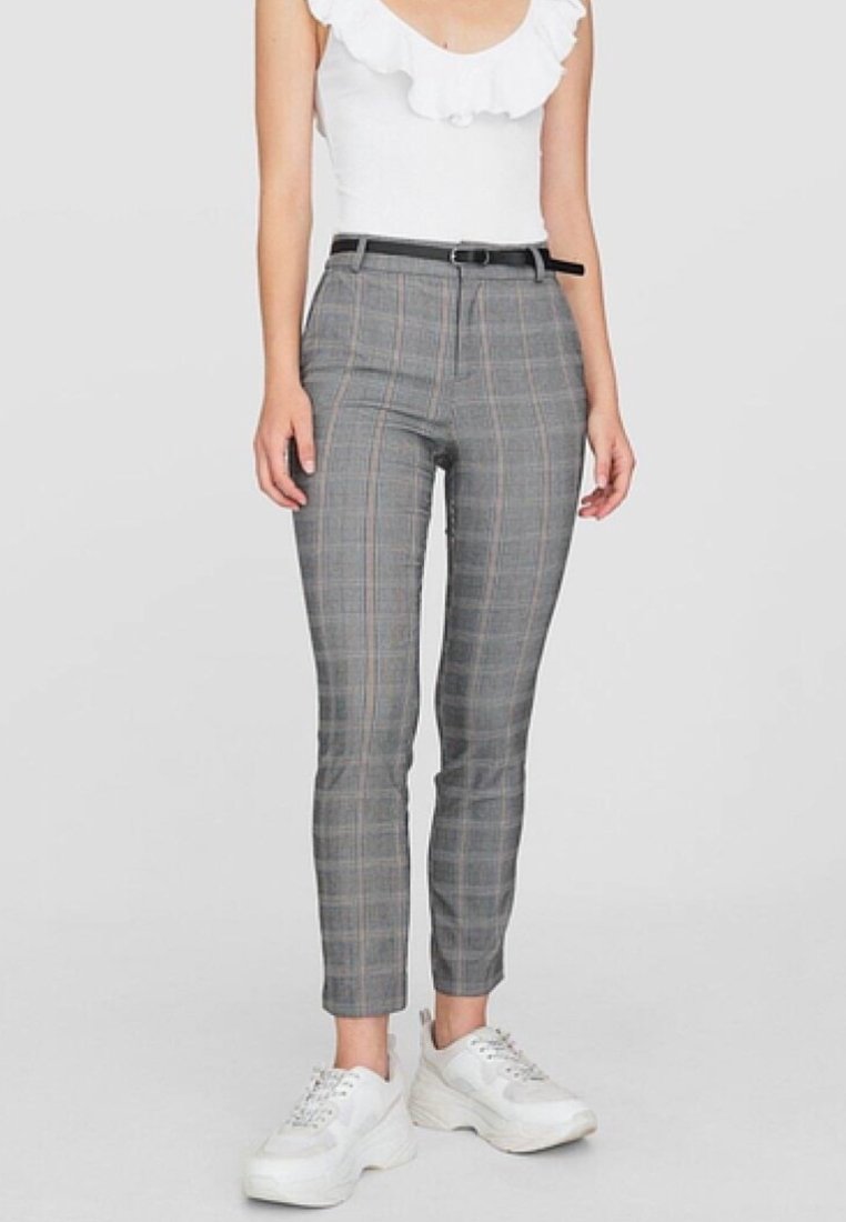 Femme portant un pantalon slim à carreaux gris avec une ceinture noire, un top blanc sans manches à volants et des baskets blanches épaisses.