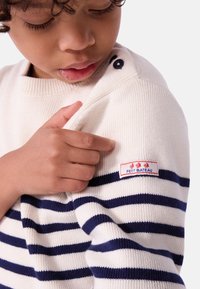 Kremgenser med marineblå horisontale striper, ribbet tekstur og knappedetaljer på skulderen. Har et Petit Bateau-logo på ermet.