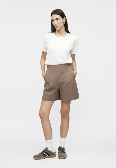 Frau mit langen dunklen Haaren, die ein weißes Kurzarmshirt, braune, strukturierte Shorts, weiße Socken und olivgrüne Sneaker trägt, steht mit den Händen in den Taschen.