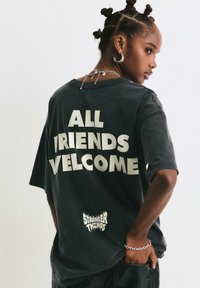 Prevelika crna majica s bijelim blok slovima: "SVI PRIJATELJI SU DOBRODOŠLI" na leđima, s malim logotipom "STRANGER THINGS" na dnu.