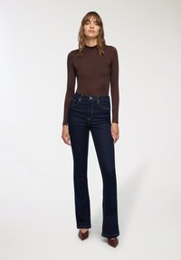 Brązowa bluzka z długim rękawem połączona z ciemnoniebieskimi dżinsami z wysokim stanem i rozszerzanymi nogawkami, ozdobionymi złotymi przeszyciami i kieszeniami. Brązowe buty na obcasie dopełniają całość.