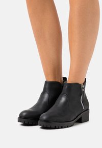 Černé kotníkové boty se bočními zipy nošené na holých nohách, s masivními podrážkami a nízkým blokovým podpatkem na bílém pozadí.
