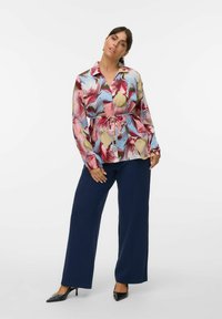 Blouse colorée avec un motif floral, manches longues et col à boutons, associée à un pantalon large navy et des talons hauts noirs.