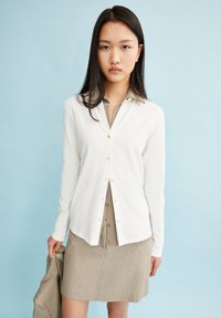 Witte blouse met lange mouwen en een knoopsluiting, met een kraag en een beige shirt eronder, gecombineerd met een beige geruite rok.