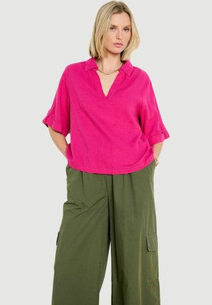 Kobieta ubrana w jasnoróżową bluzkę z kołnierzykiem i podwiniętymi rękawami oraz luźne oliwkowozielone spodnie bojówki, stoi z rękami w kieszeniach.