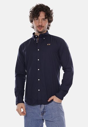 Uomo con capelli ricci che indossa una camicia blu navy abbottonata con logo ricamato e jeans azzurri, in piedi con una mano in tasca.