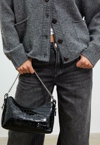 Sort patenteret læderhåndtaske med krokodillemønster, udstyret med en sølv kædrestropp. Bæres med en grå cardigan og mørke jeans.