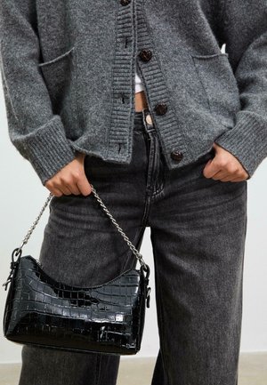 Borsa a mano in pelle verniciata nera con texture a coccodrillo, dotata di una tracolla in catena argentata. Indossata con un cardigan grigio e jeans scuri.